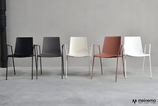 Vijf stapelbare designstoelen in een rij: zwart, donkergrijs, wit, terracotta en wit, met metalen frames op betonvloer tegen