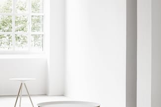 Minimalistisch interieur met witte ronde bijzettafel op driepotige houten poten, grote ramen met uitzicht op groen, en witte