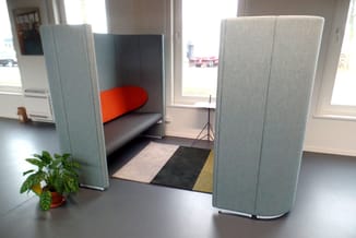 Akoestische scheidingswanden in grijs stof rond zitbank met zwart onderstel en oranje rugkussen in kantoorruimte.