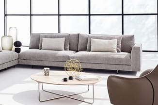 Grijze loungebank met lichte kussens, bruin fauteuil, wit ovaal tafeltje met metalen onderstel en gouden decoratieve bol in m