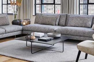 Grijze hoekbank met kussens, zwarte salontafel, lichtgrijs vloerkleed, ronde poef en grote ramen in moderne kantoorruimte.