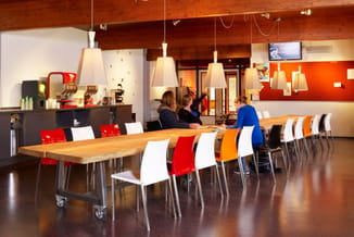 Moderne vergaderruimte met houten tafel op wielen, kleurrijke stoelen (rood, wit, oranje), hangende trapeziumvormige lampen,