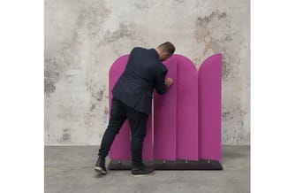 Man in donker pak buigt zich over roze akoestische scheidingswand met gebogen bovenkant tegen betonnen muur.