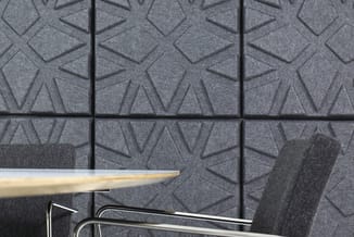 Grijze akoestische wandpanelen met driehoekig reliëfpatroon, geplaatst in rasteropstelling met zwarte voegen, met deel van bu