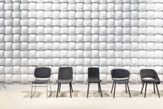 Vijf donkergrijze stoelen met zwart metalen onderstel in een rij tegen een witte tegelmuur op een beige vloer.