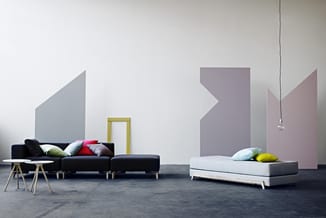 Showroom met zwarte modulaire bank met gekleurde kussens, wit bed, houten bijzettafels, geel frame en grijze/roze wandelement