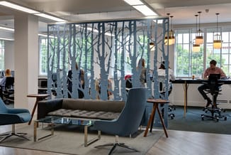 Modern kantoor met grijsblauwe bosscherm, grijze sofa, houten tafels en hangende pendels boven werkplek aan raam.