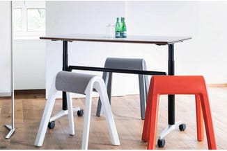 Hoog bureau met zwart stalen onderstel en grijs blad, bezet met drie gekleurde krukken op wielen: wit, grijs en rood-oranje.