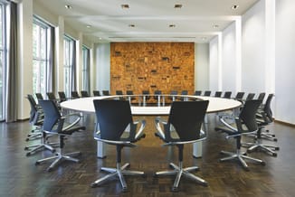Ronde vergadertafel wit met zwarte ergonomische kantoorstoe­len, houten vloer, grote ramen, gouden muurschildering met archit