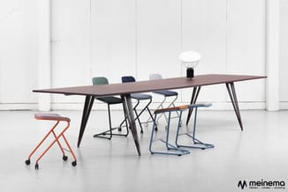 Vergadertafel met donker blad en zwart onderstel, omgeven door diverse mobile stoelen in rood, zwart, blauw en grijs. Witte i