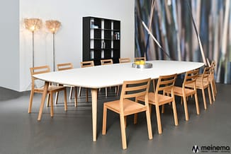 Vergadertafel met wit blad en houten onderstel, omringd door houten stoelen. Zwarte boekenkast, vloerlamp en abstract kunstwe