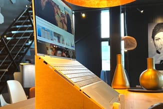 Houten laptopstandaard in natuurlijke hout-kleur met "ln.moniem" tekst, met zwarte hangende lamp met gouden binnenkant erbove