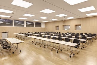 Moderne vergaderruimte met lange witte tafelbladen op wielen, zwarte kantelstoelen in rijen, lichte houten vloer, beige wandb