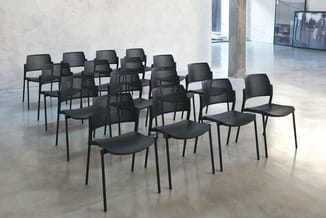 Zwarte stapelstoelen met perforeerde rugleuning en zitting, opgesteld in rijen op betonnen vloer in moderne kantoorruimte.