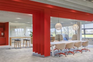 Modern kantoor met rode structurele kolommen, witte vergadertafel, beige bureaustoelen op rode onderstel, hangende bollampen