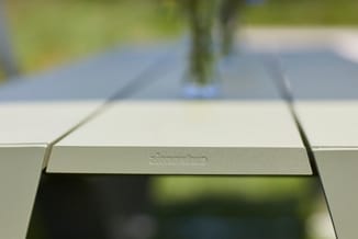 Witte tafelblad in close-up met groene buitenomgeving op achtergrond, waarschijnlijk terrastafels of tuinmeubels.