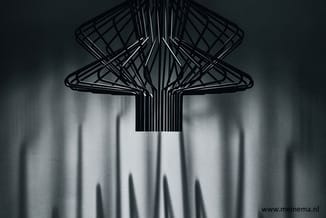 Zwarte draadwerk hanglamp met geometrische structuur, bovenaanzicht met schaduwwerping op wit oppervlak.