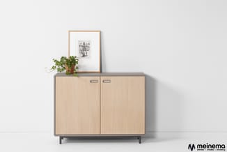 Lichte houten kast met twee deuren en metalen frame tegen witte muur, met potplant en ingelijste tekening erop.