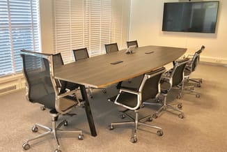 Moderne vergaderruimte met houten tafel, zwarte mesh-stoelen op wielen, wandmonteerde TV en jaloeziën aan ramen.