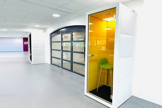 Gele belcel met glazen wand en groene stoel, in moderne kantoorruimte met witte wanden en grijze vloer.