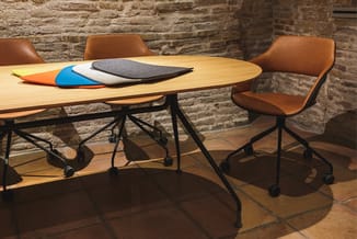 Ovale vergadertafel in licht hout met zwart frame en wielen, omgeven door bruinleren draaistoelen met zwart onderstel. Kleurr