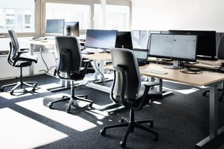 Modern kantoor met houten bureaus, ergonomische zwarte bureaustoelen, meerdere monitors en grote ramen met daglicht.