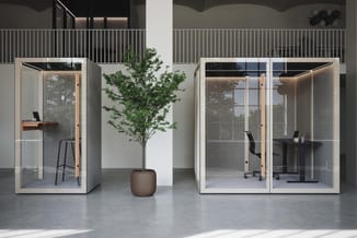 Twee witte akoestische belcellen in moderne kantooromgeving, beide voorzien van glazen wanden, werkblad en stoel. Tussen de c