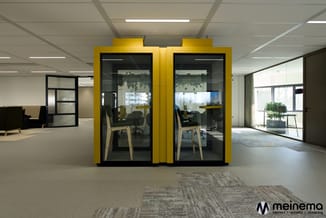 Gele akoestische belcel met glazen deuren en zwart frame in open kantoorruimte met beige vloerbekleding.