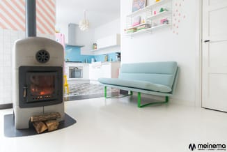 Moderne woonkamer met lichtblauwe sofa op groen metalen frame, betonnen houtkachel, witte keuken met blauwe achterwand en ope