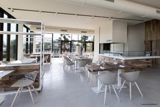 Modern restaurant-interieur met houten tafels, witte stoelen, open keuken en grote ramen met uitzicht op terras en palmen.