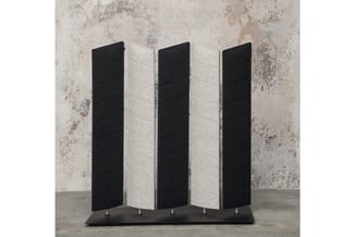 Akoestische scheidingswand met vijf panelen in zwart en grijs, op zwart voetstuk, tegen gepleisterde achtergrond.
