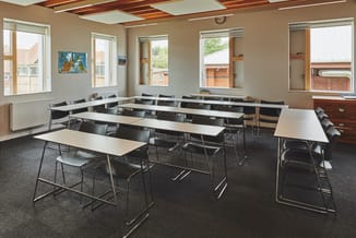 Moderne trainingsruimte met witte rechthoekige tafels, zwarte stapelstoelen en houten poten, groot aantal ramen, beige muren,