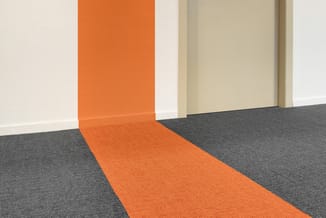 Oranje vloerbekleding loopt als pad door kantoorruimte met grijze vloer, witte en beige wanden met deuropening.