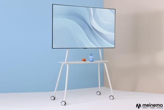 Wit kantelbaar tv-standaard op wielen met plank, dragende een monitor met blauwe achtergrond en decoratieve objecten.