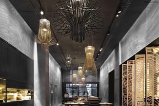 Modern restaurant met hangende gouden en zwarte design-lampen, grijze gestucte wanden, glazen scheidingswanden en houten deco