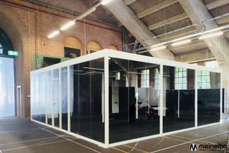 Witte belcel met zwarte glazen wanden in industrieel pand met bakstenen muur en betonnen plafond.