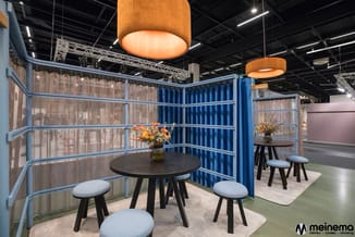 Ronde zwarte tafel met lichtblauwe stoelen in akoestische cel met blauwe gordijnen, gouden hanglamp en metalen frame.
