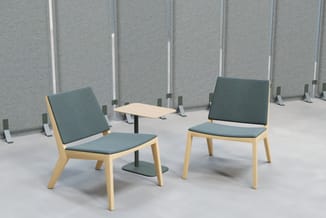 Twee houten stoelen met grijsgroene bekleding en een laag houten tafelblad in een kantoorruimte met grijze akoestische wandpa