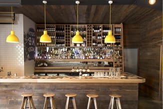 Moderne bar met houten front, gele hanglampjes, houten barkrukken, houten wandkasten met flessen, witte tegels en donker hout
