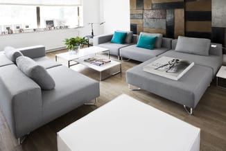 Moderne woonkamer met grijze modulaire banken, witte salontafels, turquoise kussens, houten vloer en houten wandpanelen.