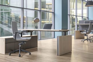 Moderne kantoorinrichting met houten bureaus, grijze ergonomische stoelen en grote ramen met uitzicht op stadsgebouwen.