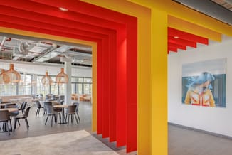Kantoorruimte met rode en gele wandelementen, houten hangende lampen, grijze stoelen rond tafels, glazen wand en kunstwerk aa