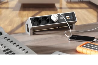 Bureaustekkerdoos wit/zwart met twee geaarde stopcontacten, USB-poorten en schakelaar op houten bureau met smartphone.