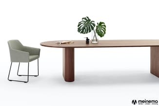 Ovale houten vergadertafel met centraal voetsteuk, groene stofstoel met zwarte poten en glazen vaas met monstera plant.