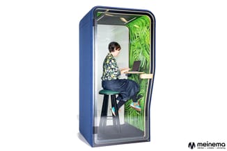 Blauwe akoestische telefooncel met glazen voorkant, waarin een persoon op een zwarte kruk aan een houten werkblad met laptop