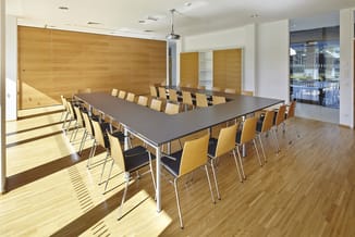 Moderne vergaderruimte met grijze rechthoekige tafel, gele stapelstoelen, houten wanden en grote ramen met zonlicht.