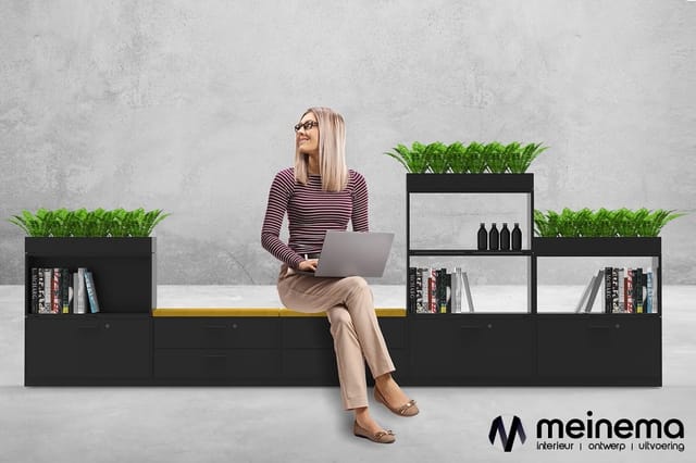 Vrouw met laptop zittend op zwarte modulaire kastencombinatie met gele accent, planters en boeken tegen betonnen muur.