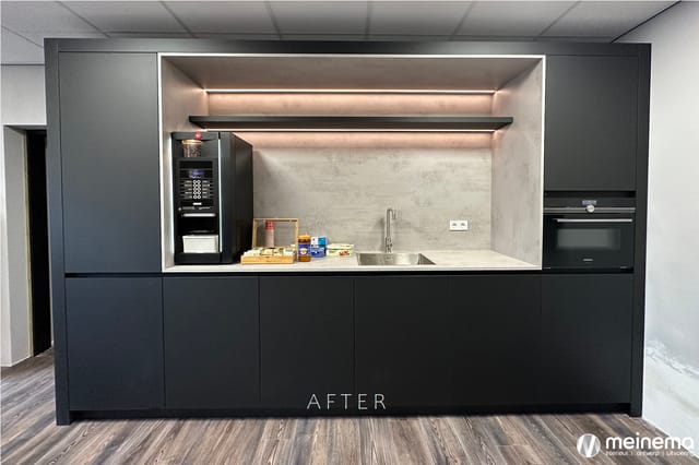 Moderne kantoorkeukeneenheid met zwart front, betonlook achterwand, ingebouwde espressomachine, oven en spoelbak. Tekst "AFTE