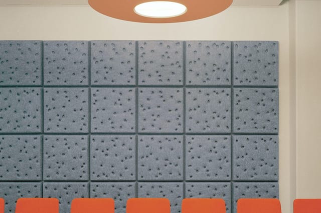 Grijze akoestische wandpanelen in 4x6 grid met donkerbruine stippen, oranje stoelen eronder, ronde plafondlamp met terracotta