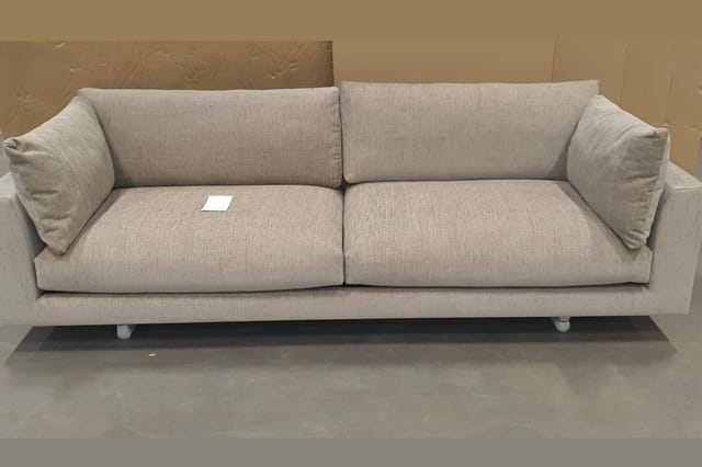 Grijsbeige linnen sofa met twee zitplaatsen, zwevend ontwerp op witte poten, twee armkussens, in tentoonstellingsruimte.